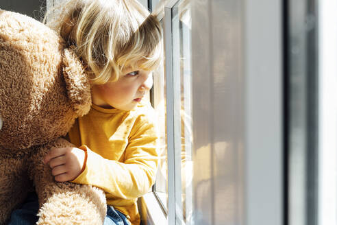 Boy with soft toy looking through window at home - VPIF06255