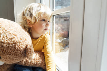 Niedlicher Junge mit Plüschtier schaut durch das Fenster zu Hause - VPIF06256