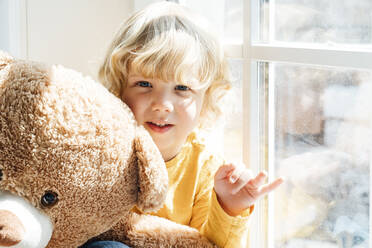 Niedlicher Junge mit Teddybär sitzt am Fenster zu Hause - VPIF06257