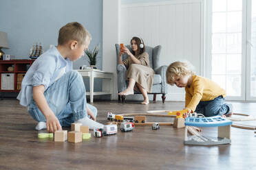 Siblings playing with toy train set while mother using smart phone sitting on armchair at home - VPIF06262