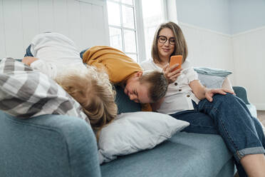 Glückliche Frau benutzt ihr Smartphone, während ihre Söhne zu Hause auf dem Sofa spielen - VPIF06286