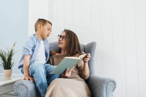 Ein Junge liest ein Buch, während seine Mutter zu Hause auf einem Sessel sitzt - VPIF06328