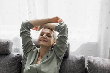 Relaxed mature woman sitting on couch in living room with closed eyes - LLUF00596