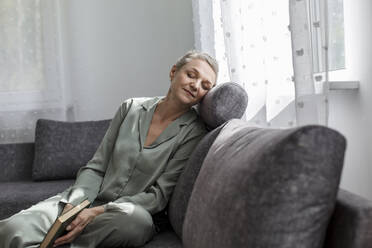 Relaxed mature woman sitting on couch in living room with closed eyes - LLUF00599