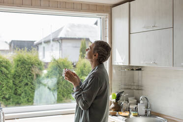 Reife Frau hält Teeglas am Fenster in der Küche - LLUF00614