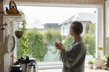 Mature woman holding tea glass at the window in kitchen - LLUF00615