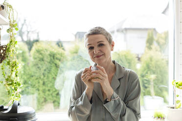 Mature woman holding tea glass at the window at home - LLUF00617