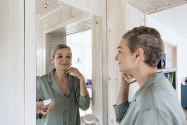 Reife Frau schaut in den Spiegel und trägt Gesichtscreme auf - LLUF00621