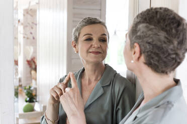 Confident mature woman looking in mirror at home - LLUF00626