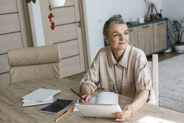 Nachdenkliche Frau sitzt zu Hause am Tisch mit digitalem Tablet und Notizbuch - LLUF00635