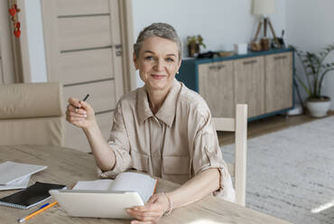 Porträt einer Frau, die ein digitales Tablet benutzt und sich am Tisch zu Hause Notizen macht - LLUF00638