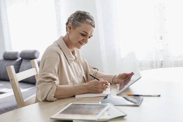 Woman using digital tablet and taking notes at table at home - LLUF00639