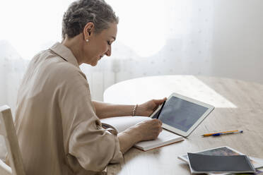 Frau hält digitales Tablet mit Daten und macht Notizen am Tisch zu Hause - LLUF00640