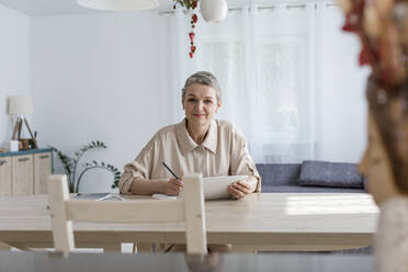 Porträt einer Frau, die ein digitales Tablet benutzt und sich am Tisch zu Hause Notizen macht - LLUF00644