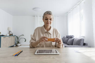 Frau sitzt zu Hause am Tisch, hält eine Karte und benutzt ein digitales Tablet - LLUF00648