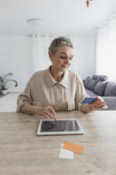 Frau sitzt zu Hause am Tisch, hält eine Karte und benutzt ein digitales Tablet - LLUF00649