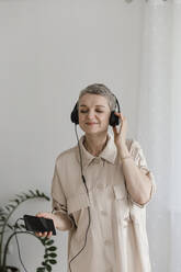 Mature woman wearing headphones listening to music at home - LLUF00665
