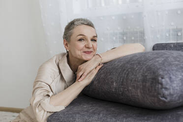 Porträt einer reifen Frau mit kurzen grauen Haaren, die sich an eine Couch lehnt - LLUF00671