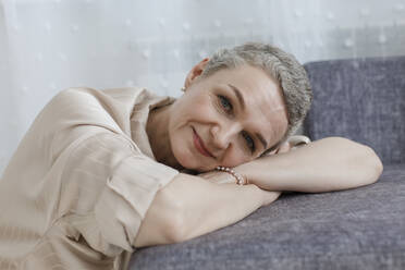 Porträt einer reifen Frau mit kurzen grauen Haaren, die sich an eine Couch lehnt - LLUF00672