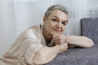 Porträt einer reifen Frau mit kurzen grauen Haaren, die sich an eine Couch lehnt - LLUF00673
