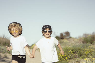 Cheerful adorable little brothers in trendy outfits holding hands while walking on sandy path amidst plants against cloudless blue sky - ADSF34842