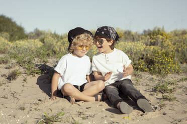 Side view of cheerful little friends in stylish clothes and sunglasses smiling happily while sitting back to back on sandy beach on sunny summer day - ADSF34845