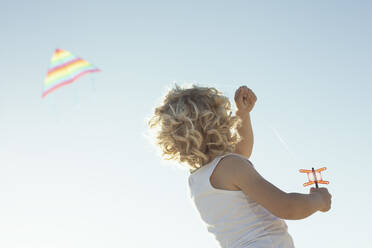 From below back view of unrecognizable cute little boy with curly blond hair launching colorful kite under cloudless blue sky on sunny summer day - ADSF34849