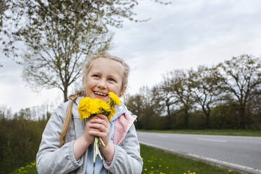 Happy girl holding bunch of dandelion flowers near road - IHF00872