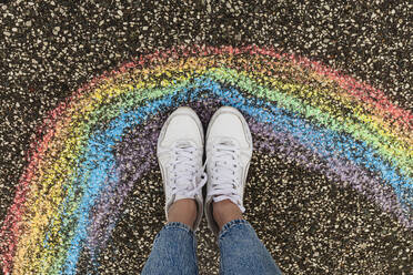 Woman wearing white shoes standing by rainbow drawing on road - IHF00881