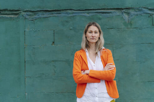 Confident woman standing with arms crossed in front of green wall - OGF01262