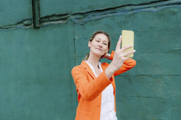 Beautiful woman taking selfie through smart phone by green wall - OGF01265