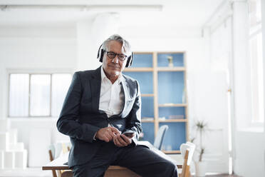 Senior businessman holding mobile phone listening music through wireless headphones in office - JOSEF10230