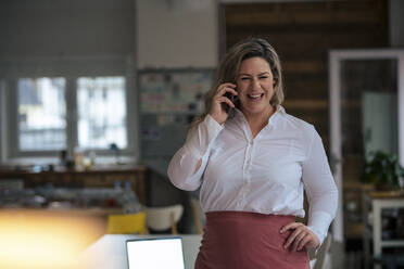 Glückliche Geschäftsfrau mit Übergröße, die im Büro mit einem Mobiltelefon spricht - JOSEF10237