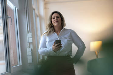 Selbstbewusste Geschäftsfrau mit Hand an der Hüfte, die ein Smartphone im Büro hält - JOSEF10256