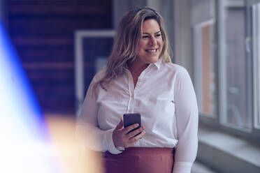 Smiling plus size businesswoman with smart phone at work place - JOSEF10262