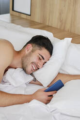 Happy man holding mobile phone waking up in bed at home - AODF00592