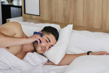 Junger Mann, der zu Hause im Bett liegt und über sein Handy spricht - AODF00593