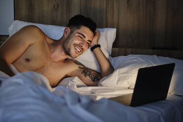 Glücklicher Mann schaut auf einen Laptop, der zu Hause im Bett liegt - AODF00601