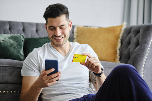 Smiling man holding credit card doing online shopping through smart phone at home - AODF00610