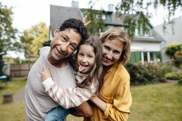 Smiling man giving piggyback ride to daughter sticking out tongue by woman in front of house - JOSEF10286