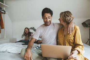 Mann mit Laptop, der neben einer Frau sitzt und seine Tochter und seinen Sohn beobachtet, die zu Hause auf dem Bett spielen - JOSEF10289
