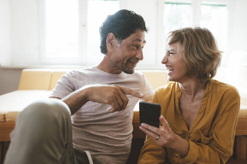Lächelnder Mann im Gespräch mit fröhlicher Frau, die ein Smartphone zu Hause hält - JOSEF10294