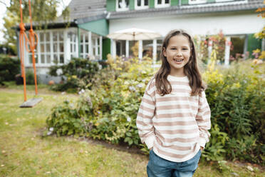 Happy girl standing with hands in pockets at back yard - JOSEF10298