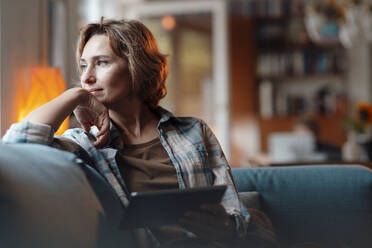 Thoughtful woman with tablet PC sitting on sofa at home - JOSEF10322