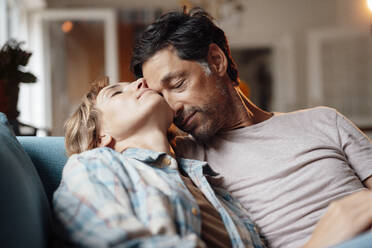 Mature man with eyes closed embracing woman sitting on sofa at home - JOSEF10327