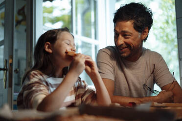 Smiling man looking at daughter eating food at home - JOSEF10340