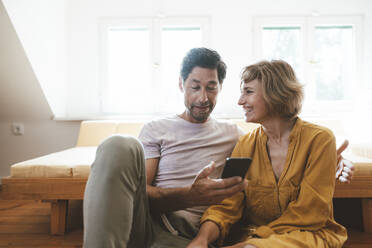 Happy woman looking at man using smart phone at home - JOSEF10356