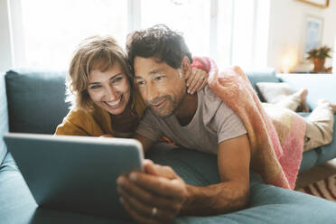 Happy mature couple using tablet PC lying on sofa at home - JOSEF10363