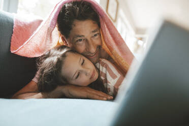 Lächelnder reifer Mann mit Tochter, die zu Hause unter einer Decke liegt - JOSEF10366