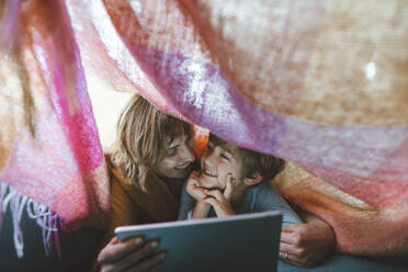 Cheerful mother and son with tablet PC lying under blanket - JOSEF10373